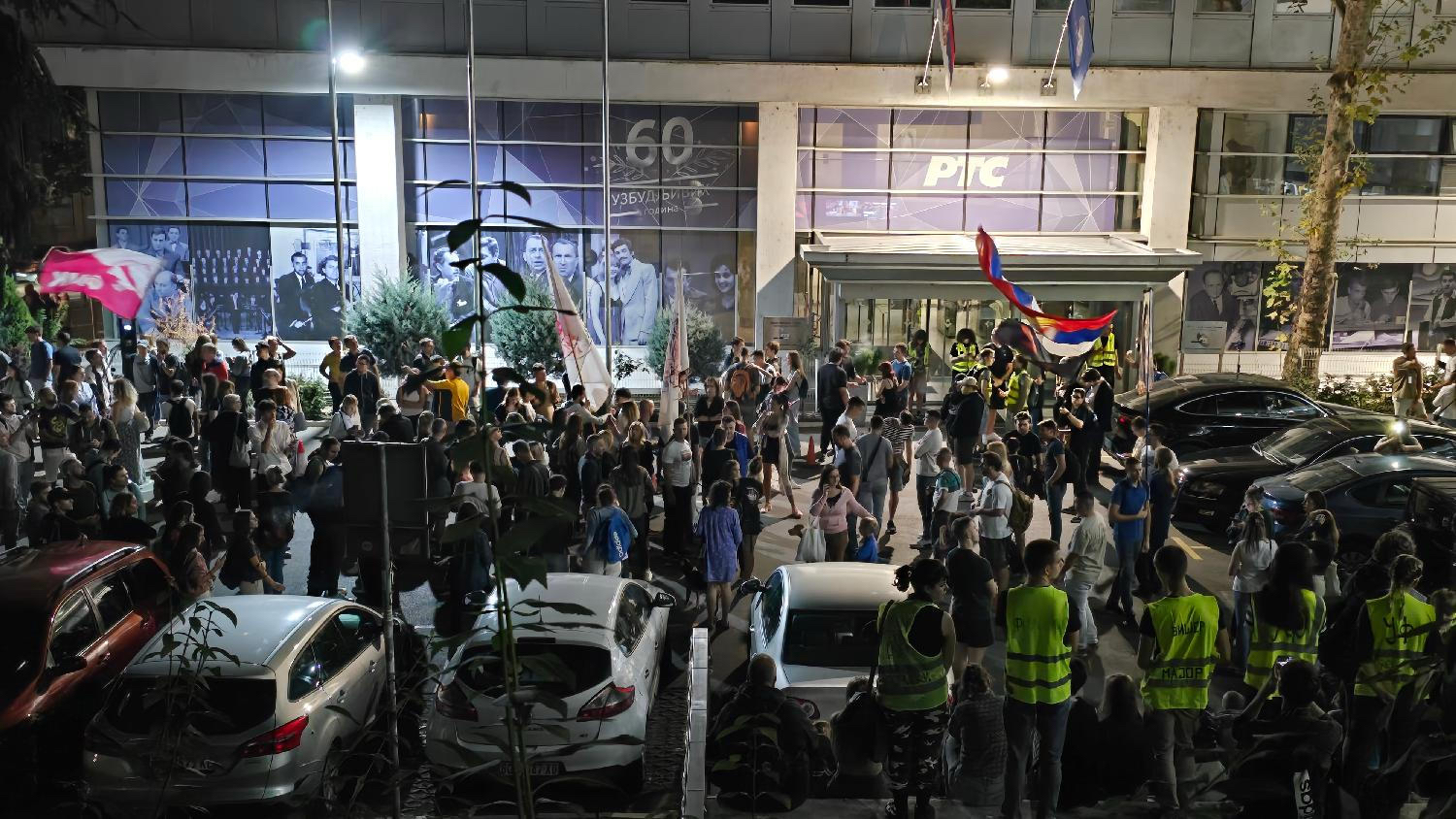 A crowd of people gathered in front of the RTS building at night, holding flags and standing near parked cars, with security personnel visible at the entrance